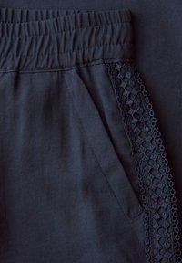 Gros plan sur un pantalon bleu marine avec une ceinture élastique, une poche latérale et un galon en dentelle décorative le long du bord de la poche.