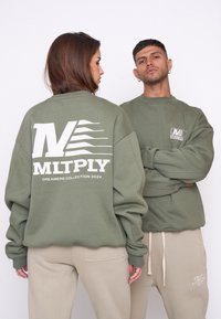 Olijfgroene sweatshirts met grote witte logo's op de achterkant en voorkant, gecombineerd met beige broeken; relaxte pasvorm en ribgebreide manchetten.