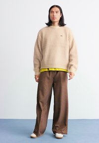 Pulôver beige de malha com uma textura canelada, usado por cima de uma camisa amarela. Calças largas xadrez castanhas e sapatos brancos completam o conjunto.