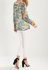 Blomsterlangærmet bluse i blå, gul og grøn nuancer, parret med hvide slim-fit bukser og sorte block-heeled sandaler.