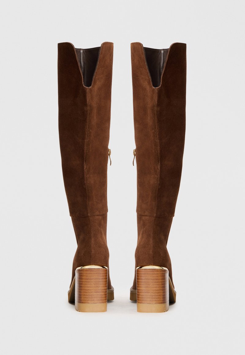 Botas de ante marrón hasta la rodilla con una abertura en forma de V en la parte superior y un tacón cuadrado, que presentan herrajes en tono dorado y una base de madera texturizada.