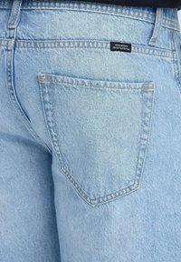 Πίσω τσέπη ανοιχτό μπλε τζιν με ραφές και μικρή μαύρη ετικέτα με την επιγραφή «DEDICATED TO DENIM PERFECTION».