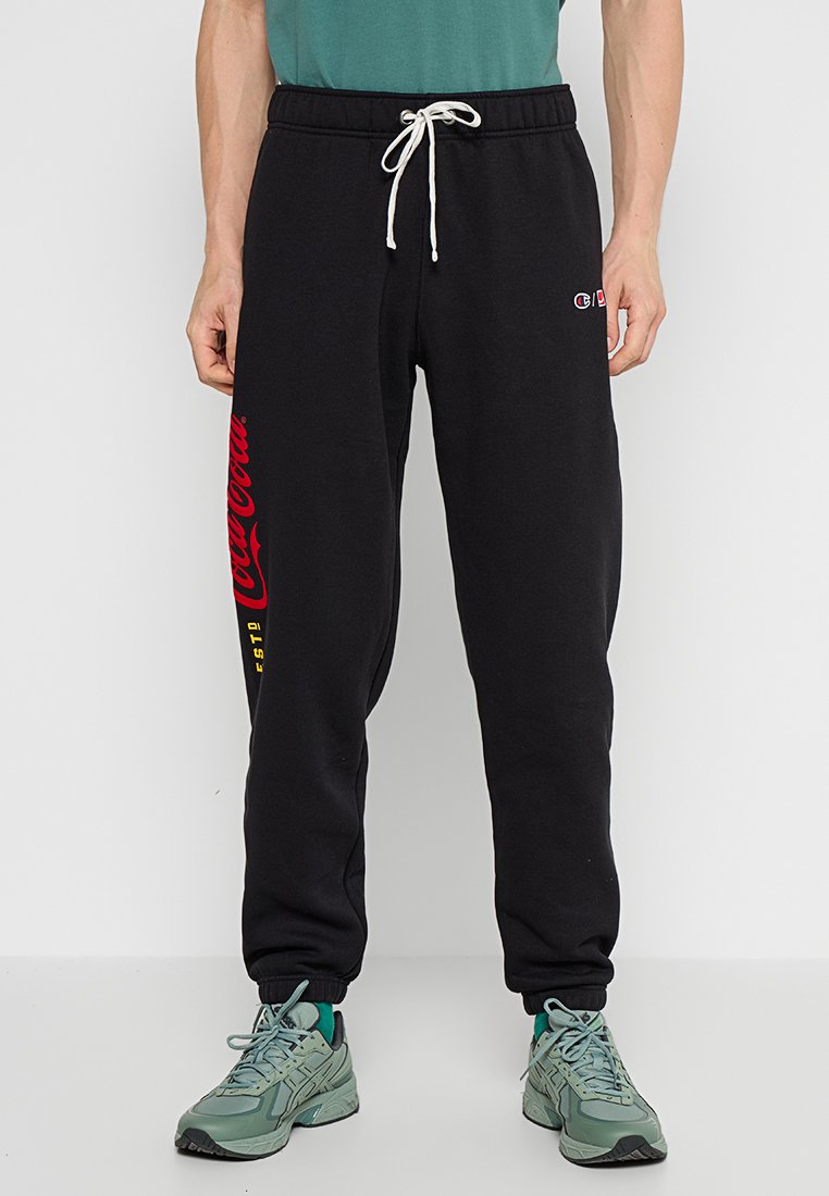 Pantaloni sportivi neri con coulisse bianca, testo rosso della Coca-Cola sulla gamba sinistra, piccolo logo Champion sulla coscia destra, indossati con sneakers verdi.