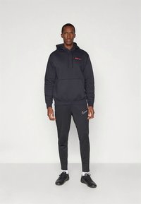 Felpa nera con cappuccio e logo rosso, abbinata a pantaloni neri a sigaretta con una fascia Nike bianca. Modello con scarpe da ginnastica nere.