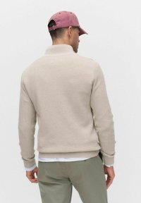 Beige stickad tröja med ett texturerat mönster, med hög krage och ribbade ärmmuddar. Modellen har på sig en rosa keps och olivgröna byxor.