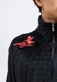 Nærbillede af sort lynlåstrøje med gentaget "AFC" mønster og rød broderet kerub med kanon på den øvre brystdel.