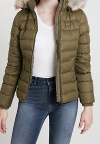 Chaqueta acolchada verde con cremallera frontal y capucha desmontable forrada de piel. Presenta un diseño acolchado y puños de canalé. Combinada con jeans azul oscuro.