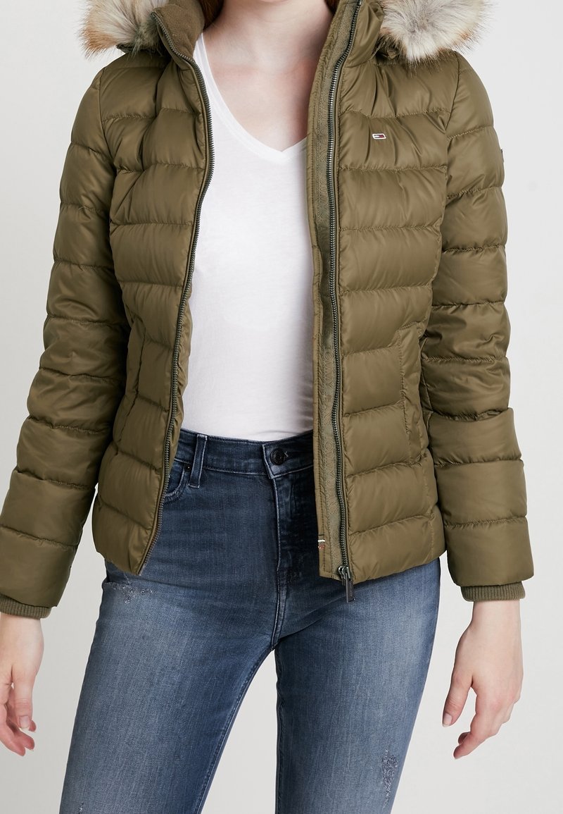 Chaqueta acolchada verde con cremallera frontal y capucha desmontable forrada de piel. Presenta un diseño acolchado y puños de canalé. Combinada con jeans azul oscuro.