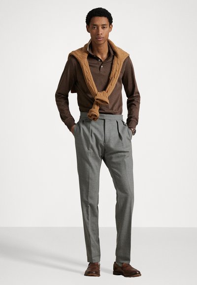 Bruine poloshirt met lange mouwen, met een kabelgebreid beige sweater om de nek geknoopt, gecombineerd met grijze geplooide broeken en bruine leren schoenen.