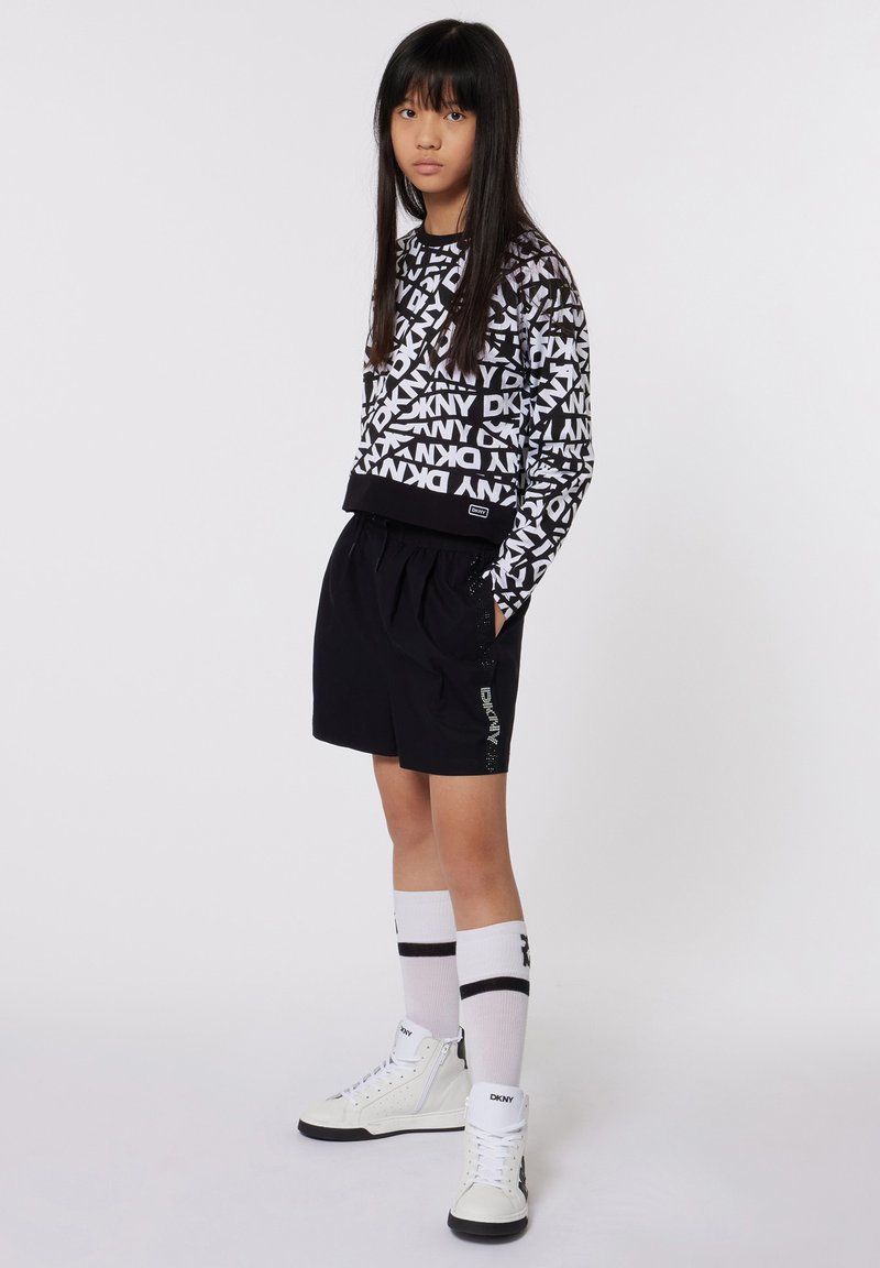 Sweatshirt noir et blanc avec un motif de logo répété, associé à un short noir avec des poches latérales. Baskets montantes blanches et chaussettes rayées.