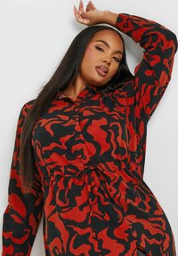 Camicia a maniche lunghe nera e rossa, con un motivo astratto ondulato, chiusura a bottoni e vita regolabile con cordino per una vestibilità adattabile.