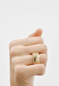 Anillo dorado con diseño apilado y estriado, que presenta secciones alternas suaves y texturizadas, llevado en una mano con los dedos curvados.