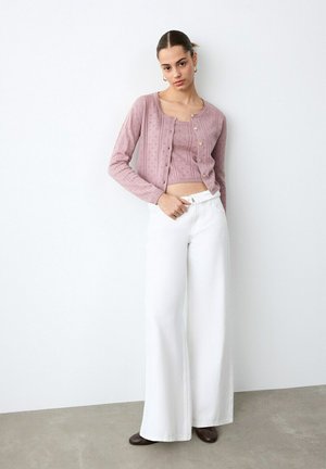 Žena v vínovom pletenom crop tope s rovnakým kardigánom, biele široké nohavice a tmavé topánky, stojaca pri jednoduchej bielej stene.