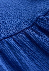 Niebieska tkanina o fakturze z pofałdowaną powierzchnią, z wyraźnymi zmarszczkami i fałdami, charakterystyczna dla lekkiego, zwiewnego materiału.