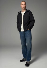 Giacca leggera nera con tasca con zip, indossata sopra una maglietta bianca e jeans blu. Scarpe da ginnastica nere completano l'outfit.