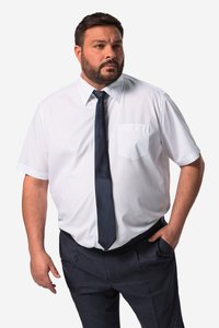 Mann mit kurzem dunklem Haar und Bart, trägt ein weißes kurzärmliges Hemd und eine dunkle Krawatte, steht mit einer Hand in der Tasche und schaut nach rechts.