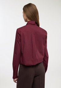 Camicia bordeaux a maniche lunghe con righe bianche verticali, presenta un colletto stretto e polsini con bottoni, realizzata in tessuto liscio, con una vestibilità rilassata.