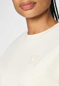 Helle beige Sweatshirt mit Rundhalsausschnitt, das ein kleines, gesticktes Logo-Patch auf der Brust in Hellgrau zeigt. Weiche Stofftextur.