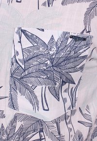 Valkoinen kangas, jossa on tummansiniset trooppiset lehtikuviot, ja siihen kuuluu sopiva kuviollinen tasku, jossa on pieni metallinen brändilogo yläreunassa.