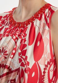 Top sans manches présentant un motif floral rouge et crème, avec un col perlé et un tissu doux se drapant élégamment.