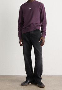 Fialová mikina s bílým logem "BOSS", v kombinaci s černými džíny a černými teniskami, stojící před jednobarevným pozadím.