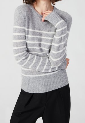 Person iført en grå sweater med hvide vandrette striber og sorte bukser, arme krydset med den ene hånd tæt på deres ansigt.