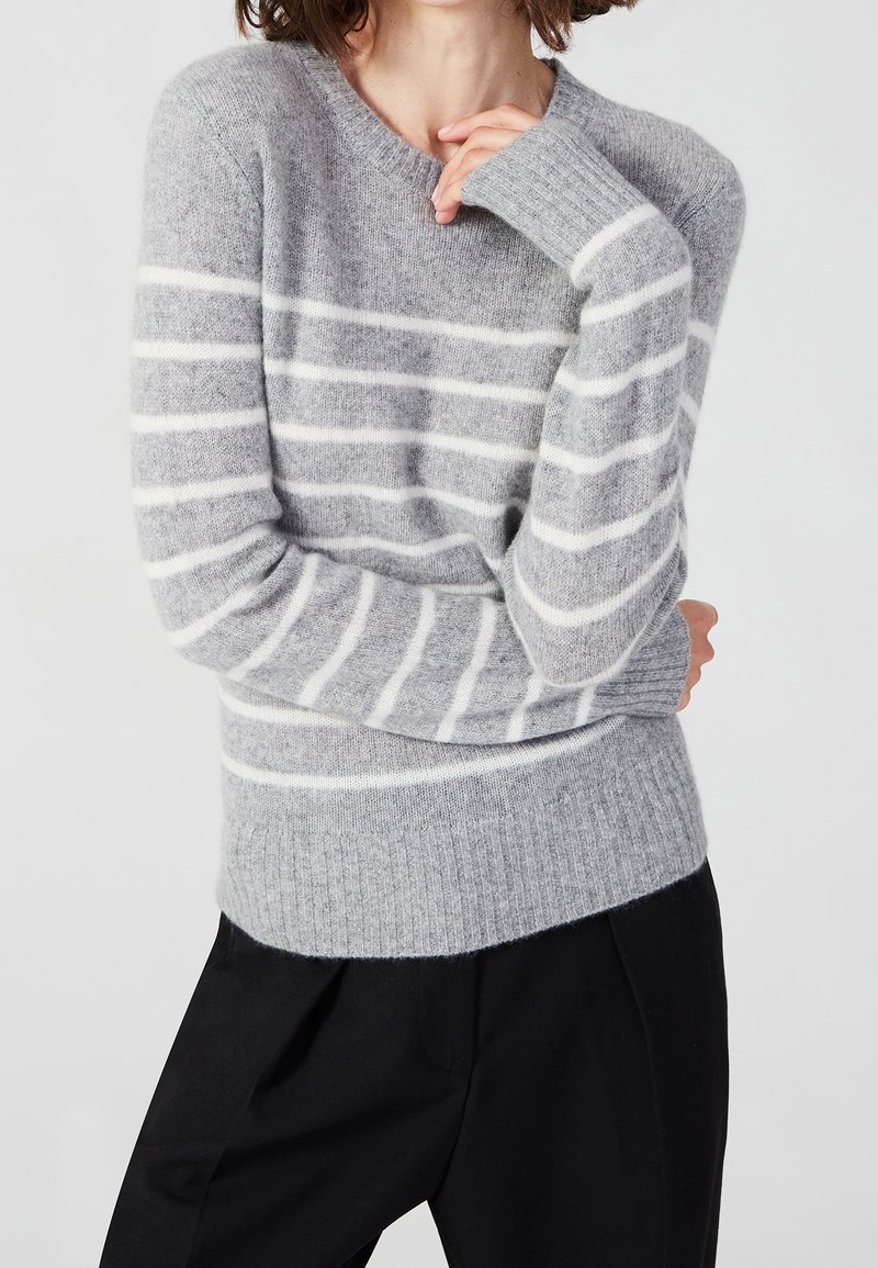 Personne portant un pull gris avec des rayures horizontales blanches et un pantalon noir, les bras croisés avec une main près du visage.