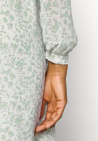 Vestido floral em verde claro e branco com um tecido texturado e transparente. Apresenta mangas bufantes e um detalhe de punho com um único botão.