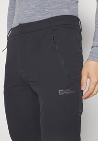 Mustat ulkoiluhousut sileällä pinnalla, joissa on vetoketjullinen tasku ja logo lahkeessa. Suunniteltu mukavuutta ja toimivuutta varten.