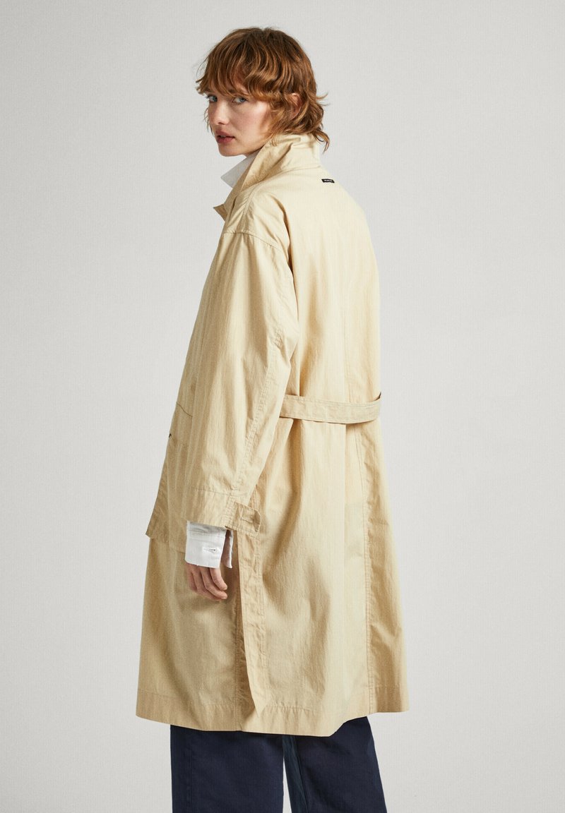 Trench-coat beige avec un col haut, coupe décontractée, fentes latérales et ceinture. Fabriqué à partir d'un tissu léger au toucher lisse.