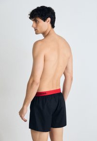 Maillots de bain noirs avec une ceinture élastique rouge portant le nom de la marque "HUGO" en noir. Le tissu est léger et doux.