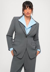 Woman with dark hair in a grey suit with side lace-up details, hands in pockets, wearing a light blue shirt with wide collar against a plain background.
