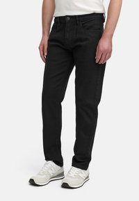 Jeans de mezclilla negra con un ajuste slim, con un diseño de cinco bolsillos, cierre de botón y detalles de costura sutiles. Llevados con zapatillas de color claro.