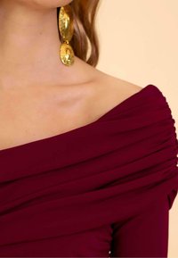 Vestido burdeos con hombros al descubierto y tejido recogido para dar textura. Grandes pendientes de oro con un patrón en relieve completan el look.