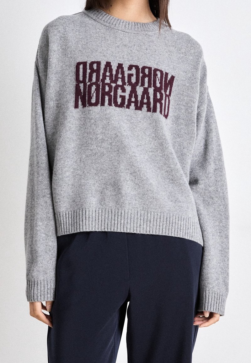 Maglione grigio lavorato a maglia con orlo e polsini a coste, con il testo "Norgaad" in un bordeaux scuro sulla parte anteriore. Texture morbida e vestibilità rilassata.