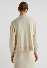 Beige strikket sweater med ribbet krave og kant, der har lange ærmer og en afslappet pasform. Glat tekstur og minimal udsmykning bagpå.