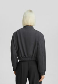 Kort svart jacka med elastisk midja, långa ärmar och en subtil texturerad finish, med ståkrage och utan synliga knäppningar.