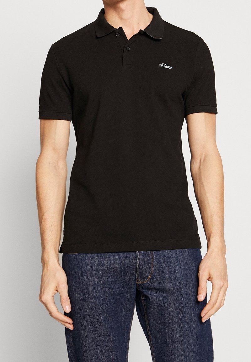 Uomo che indossa una polo nera attillata a maniche corte con colletto e bottoni, abbinata a jeans in denim blu scuro.