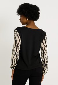 Camiseta negra con mangas largas de estampado de cebra; tela suave; corte relajado; escote en forma de barco. Pendientes de aro dorados como acento. Pantalones negros visibles.