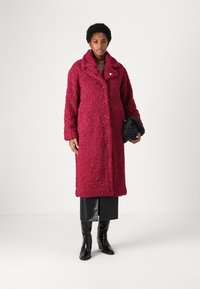 Abrigo largo burdeos, texturizado, con un gran cuello y cierre de botón; combinado con un bolso oscuro y botas de cuero negro elegantes.