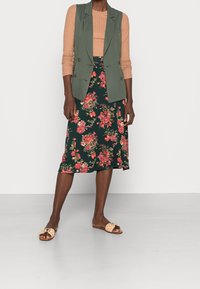 Blazer verde senza maniche sopra una maglietta color nocciola, abbinato a una gonna midi floreale verde e rossa. Sandali beige completano l'outfit.