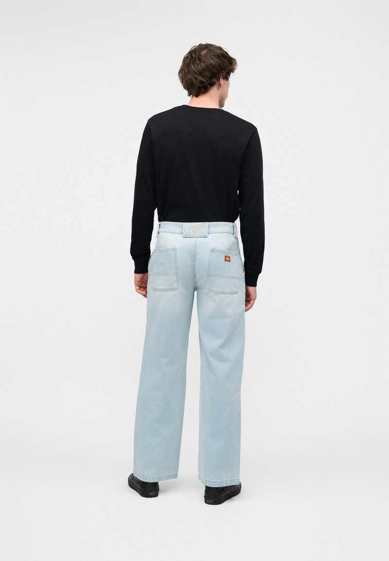 Mann mit kurzen lockigen Haaren steht mit dem Rücken zur Kamera, trägt ein schwarzes Langarmshirt, hellblaue Jeans mit weitem Bein und schwarze Schuhe vor weißem Hintergrund.