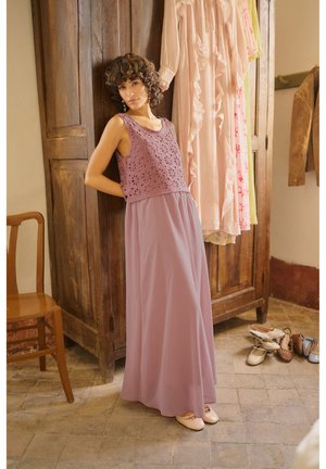 Naine roosas varrukateta heegeldatud topis ja pikas seelikus toetub puitkappile, kus ripuvad kleidid ja põrandal on jalanõud.