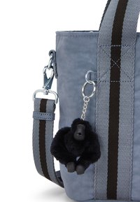 Sininen kangaskassi, jossa on teksturoitu pinta, varustettuna irrotettavalla mustalla gorillakeychainilla. Olkahihnassa on ruskeita ja mustia raitoja.