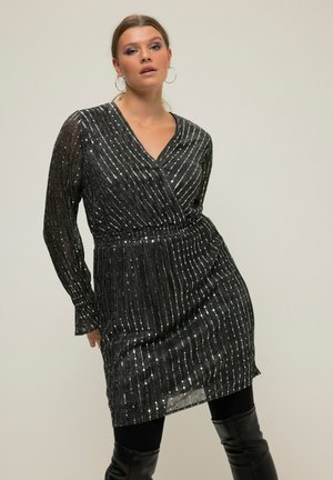 Vestido negro con mangas largas, escote en V profundo y rayas metálicas brillantes. Cintura elástica y falda fluida; combinado con leggings ajustados negros.