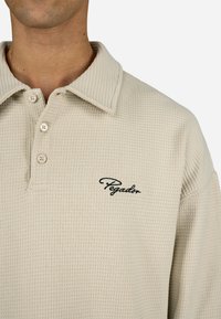 Jasno beżowa koszulka polo z dzianiny waffle z klasycznym kołnierzykiem, trzyguzkowym zapięciem i czarnym haftowanym logo "Pegadon" po lewej stronie klatki piersiowej.