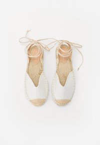 Weiße Espadrilles mit offener V-Form, geflochtener Jutesohle und dünnen beige Schnürsenkeln. Texturiertes Obermaterial mit kleinen Ausschnitten.