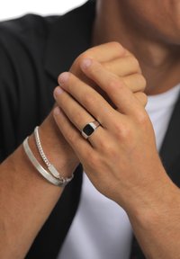 Silver ring with a hexagonal black stone, worn on a hand with light skin. Accompanied by a silver chain bracelet and a flat cuff.