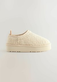 Zapatillas color crema con una textura suave, punta redondeada y una pequeña lengüeta en la parte posterior. Suela de goma gruesa en color beige con un patrón ondulado.