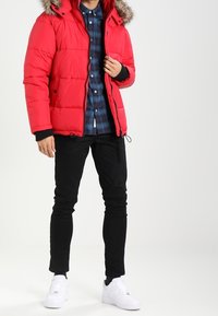 Czerwona kurtka puchowa z futrzanym kapturem, czarnymi akcentami, zapinana na zamek, w zestawieniu z niebieską koszulą w kratę, czarnymi spodniami i białymi sneakersami.
